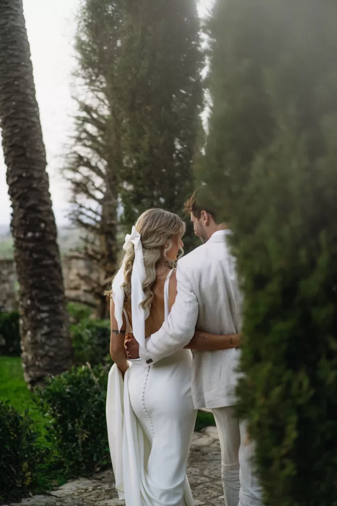 The bride in a sleek second dress with a long bow walks arm in arm with the groom through the gardens of Finca Son Collell.