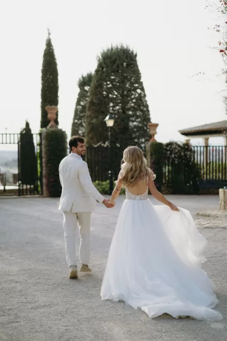 The bride and groom walk hand in hand along the tree-lined driveway at Finca Son Collell, her dress flowing behind.