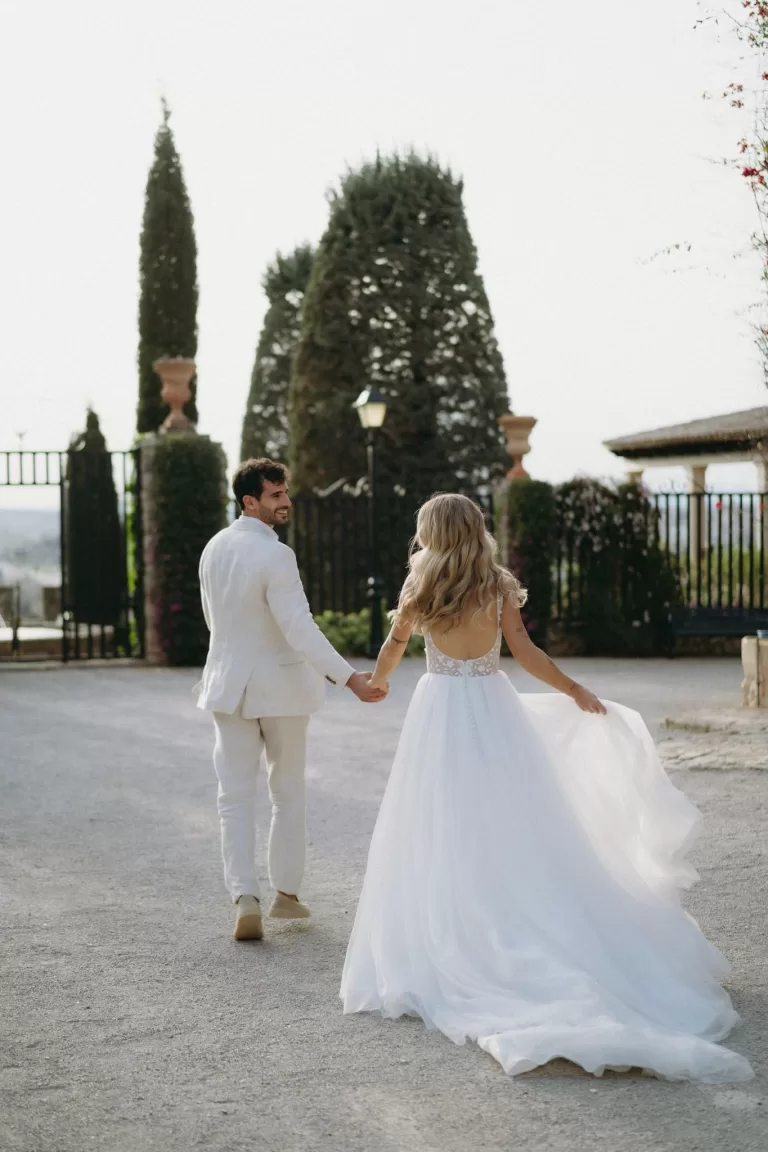 The bride and groom walk hand in hand along the tree-lined driveway at Finca Son Collell, her dress flowing behind.