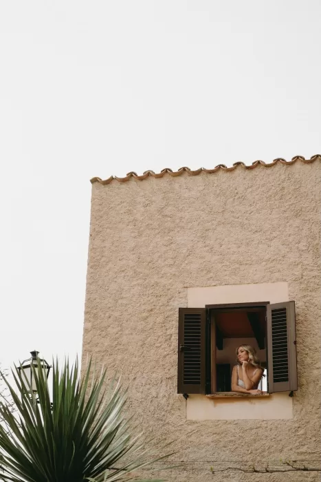 The bride gazes thoughtfully out a shuttered window of Finca Son Collell, framed by textured stone walls.