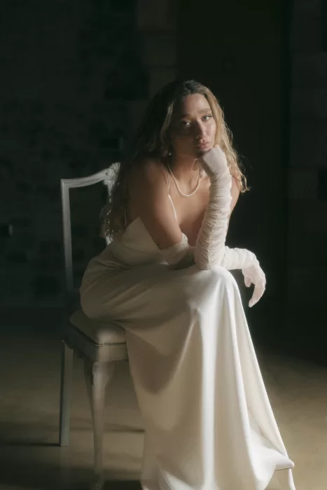 Bride in satin gown and sheer gloves sits pensively on vintage chair at Mas Torroella.