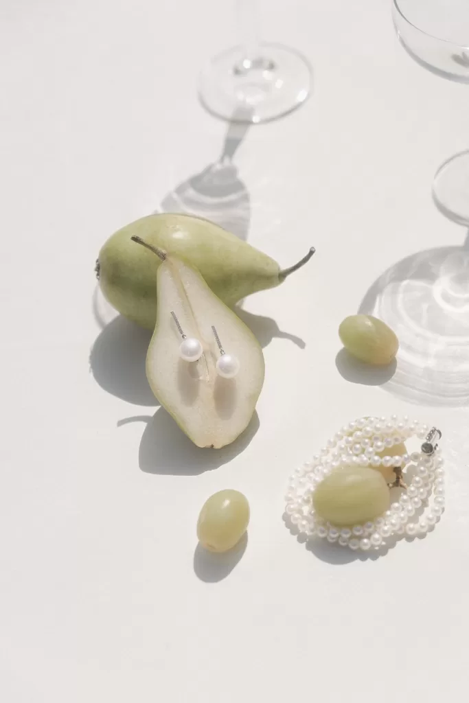 Pearl earrings displayed on sliced pear with grapes and elegant table shadows.
