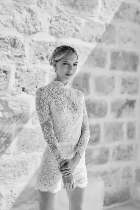 Bride in elegant lace romper poses against sunlit stone wall at Masseria Calderisi
