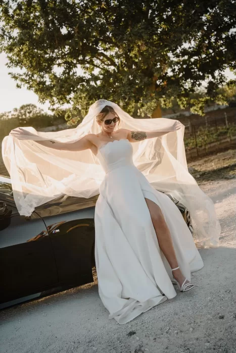 The bride twirls joyfully, lifting her veil in the golden afternoon light beside a black convertible at Masseria Monitilli.