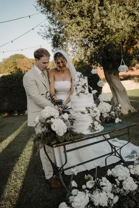 Joyful moment as the bride and groom cut their floral-adorned wedding cake under olive trees at Masseria Monitilli.