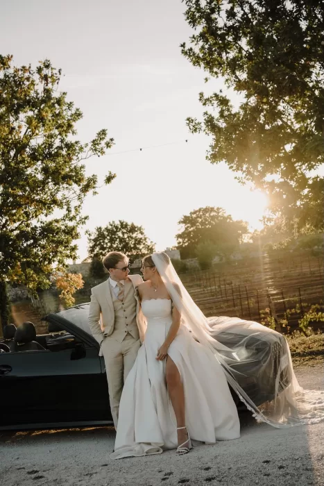 Newlyweds leaning against a convertible at sunset in the vineyard-lined driveway of Masseria Monitilli.