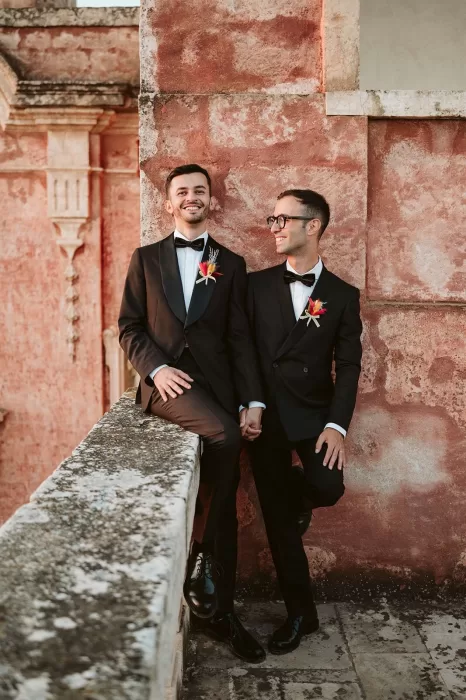 Joyful grooms holding hands on a rustic red balcony at Masseria Spina.