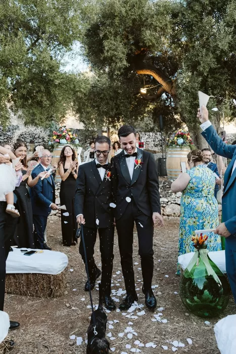 Grooms walk joyfully down the aisle with their dog as guests throw flower petals at Masseria Spina.