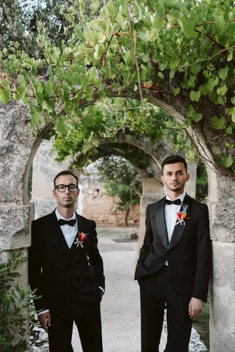 Grooms pose beneath a stone archway covered in greenery in the gardens of Masseria Spina.