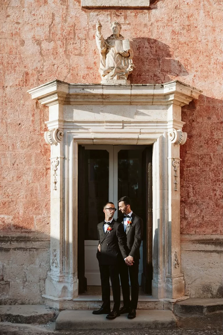 Grooms holding hands in front of a grand stone doorway at Masseria Spina, bathed in golden light