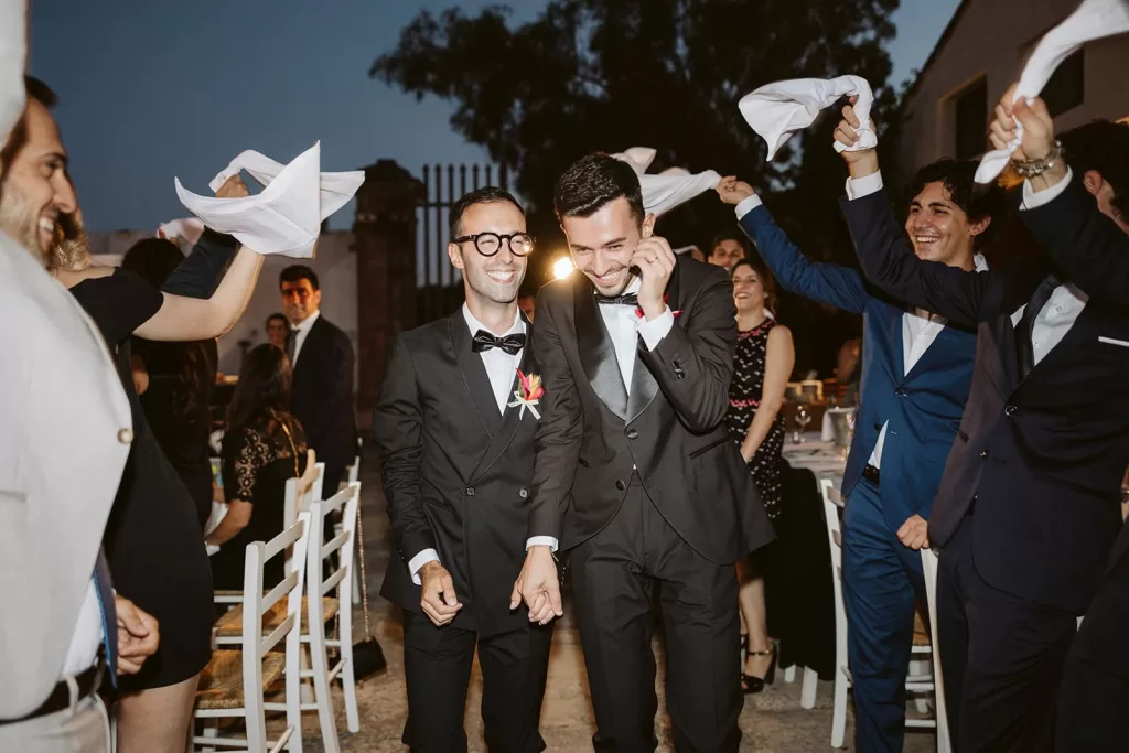Just married, two grooms walk hand in hand down the aisle at Masseria Spina, smiling as guests celebrate around them.