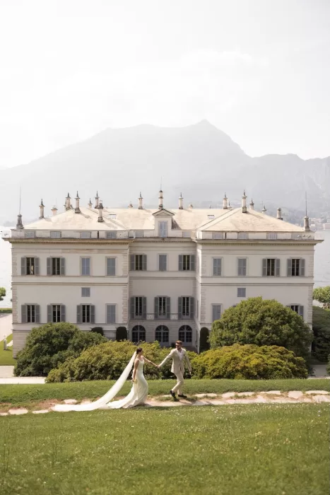 Bride and groom walking hand in hand on the lawn of Parco Civico Teresio, with the majestic villa and mountain backdrop behind them.