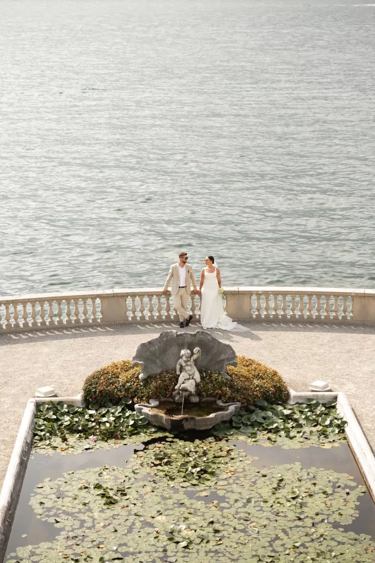 Couple walking hand-in-hand beside a lily pond and cherub fountain with Lake Como as the backdrop at Parco Civico Teresio.