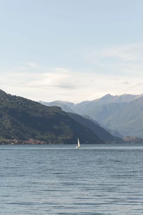 Serene lake view from Parco Civico Teresio with a single white sailboat and distant mountains under a soft blue sky.