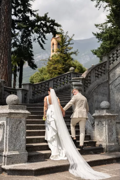 Couple walking up the grand stone staircase at Parco Civico Teresio, with a bell tower peeking through the trees behind them.