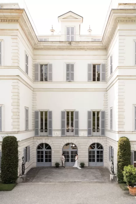 Wide view of the elegant villa at Parco Civico Teresio with the couple standing in the central courtyard, framed by tall shuttered windows.