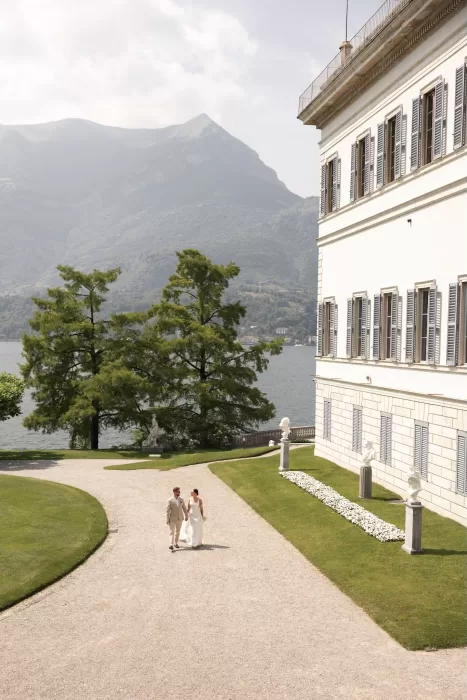 Bride and groom walking hand-in-hand along the elegant gravel path outside the villa at Parco Civico Teresio, with lake and mountains in view.