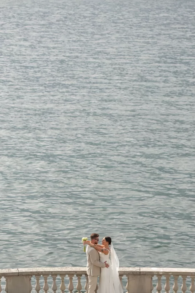 Bride and groom embracing by the lake’s edge at Parco Civico Teresio with sunlight shimmering on the water.