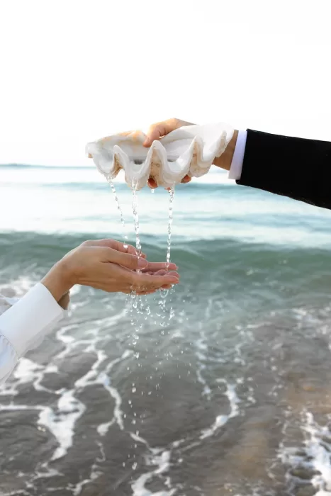 Water flows from a large seashell into the bride's hands as the waves roll in behind them—an intimate beach ritual.