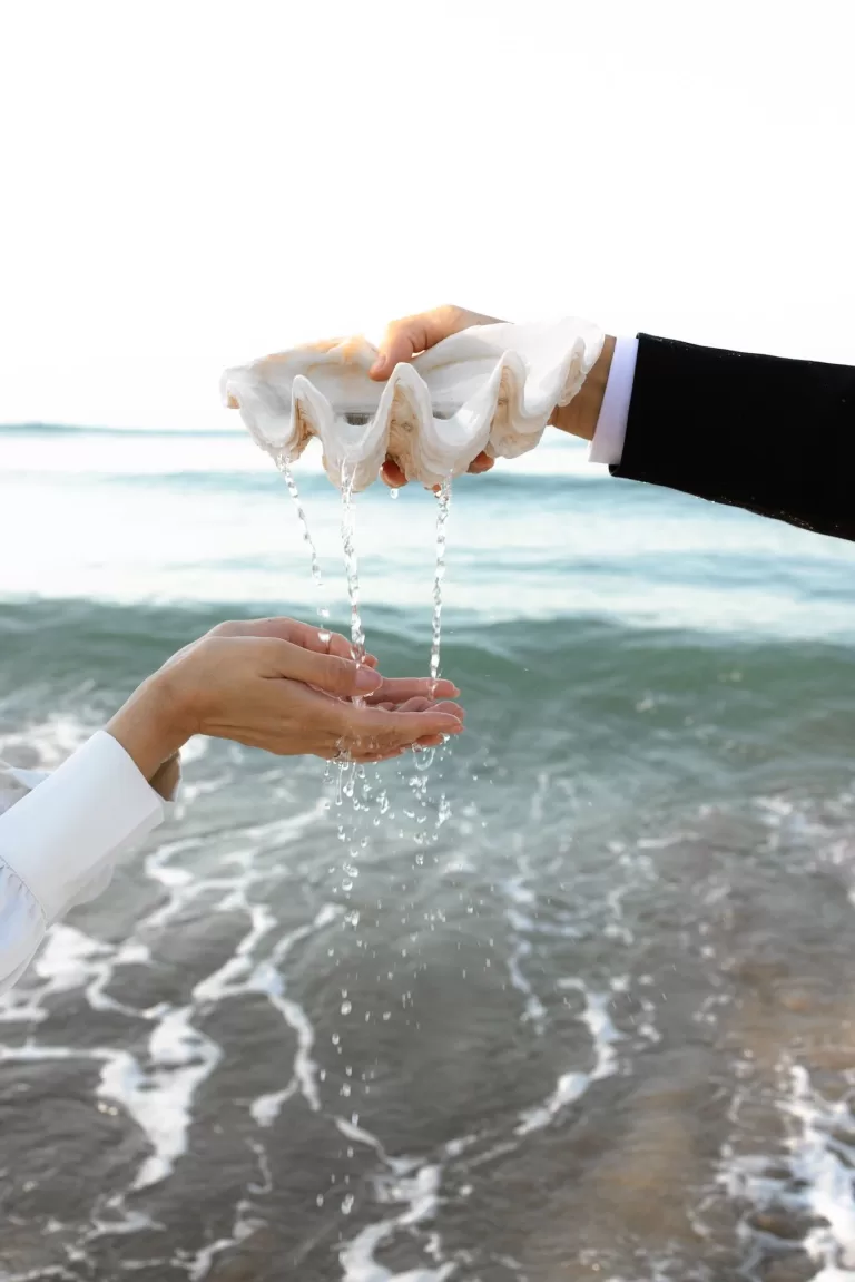 Water flows from a large seashell into the bride's hands as the waves roll in behind them—an intimate beach ritual.