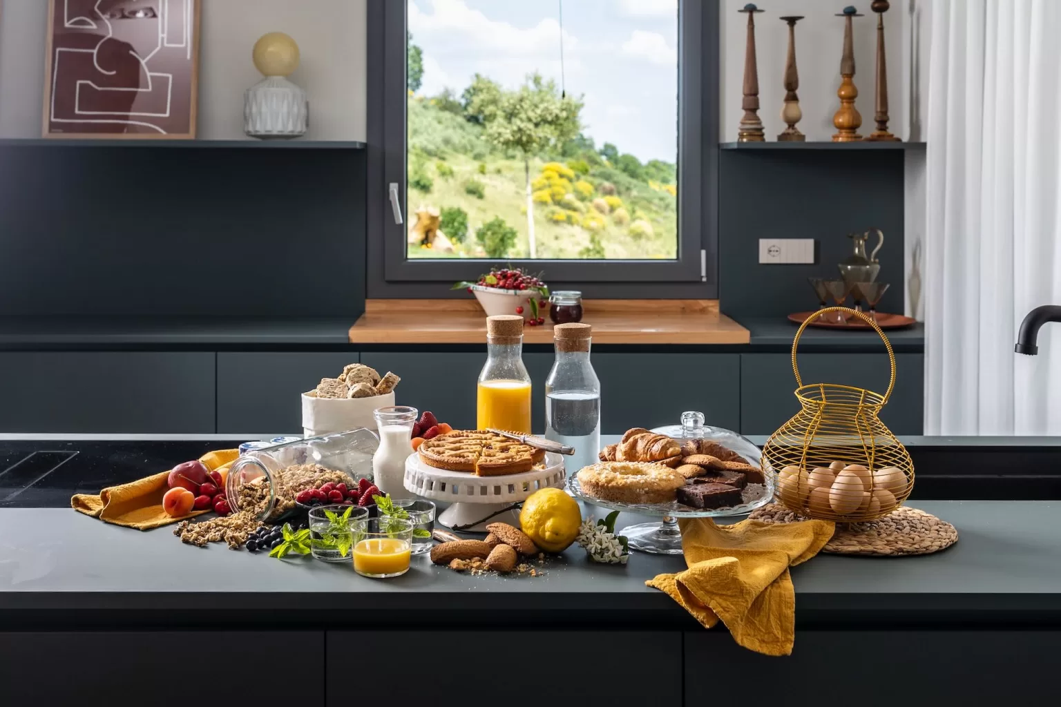A colourful breakfast spread with fresh pastries, fruit, and juice set in a modern kitchen at Villa Agavi with views of the hills.