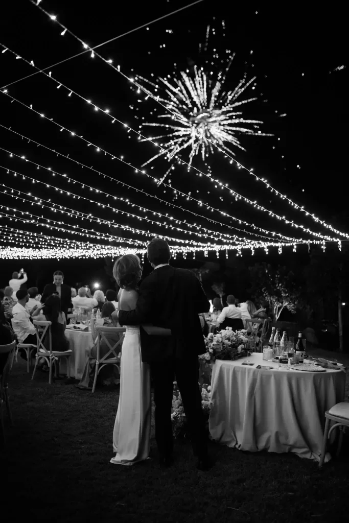 A couple embraces under a canopy of lights as fireworks light up the night sky at Villa Agavi.