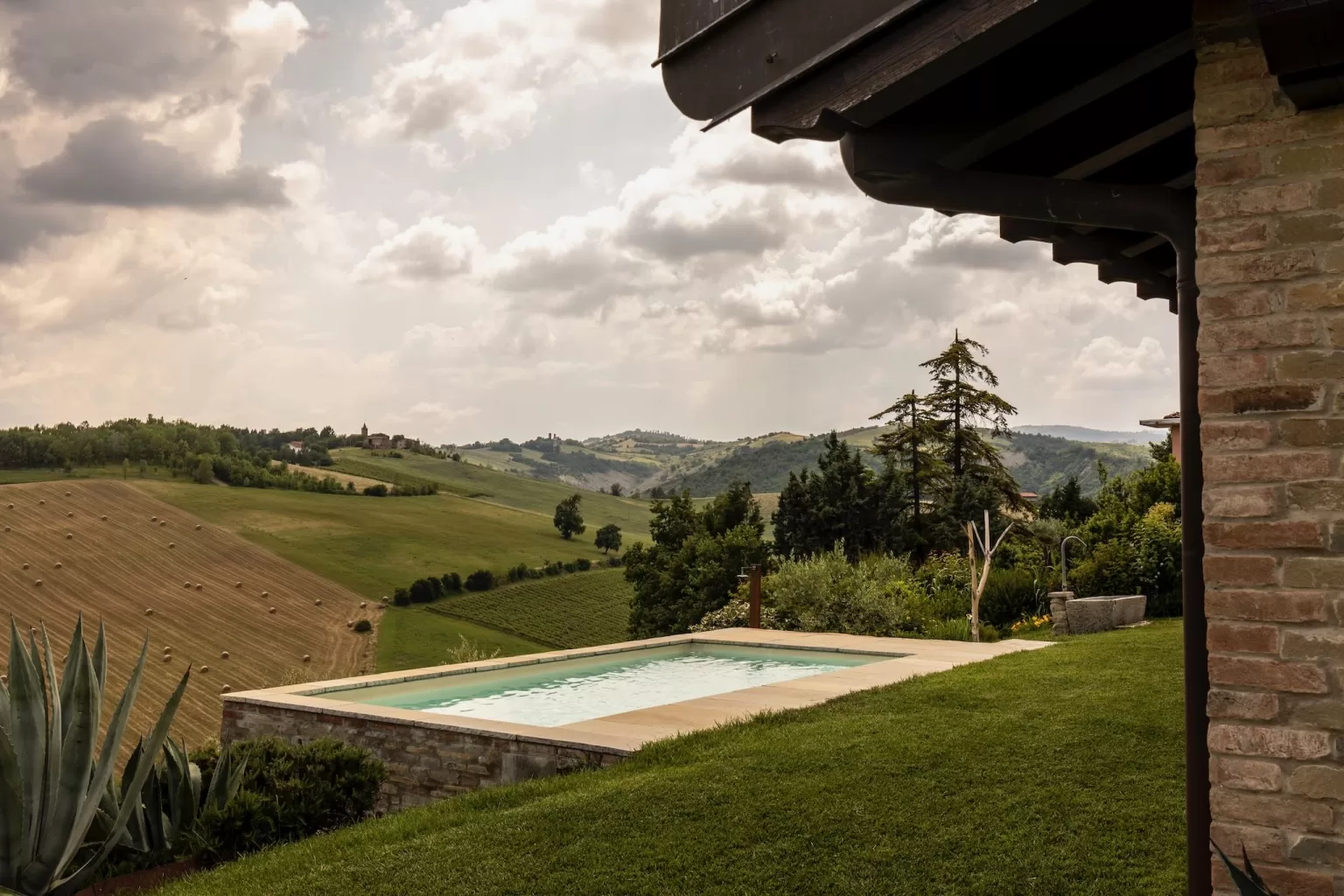 A tranquil pool at Villa Agavi opens up to wide green fields and distant hills under a softly clouded sky.