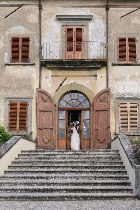 A bride steps through grand wooden doors onto the stone staircase of Villa Medicea di Lilliano’s historic facade.