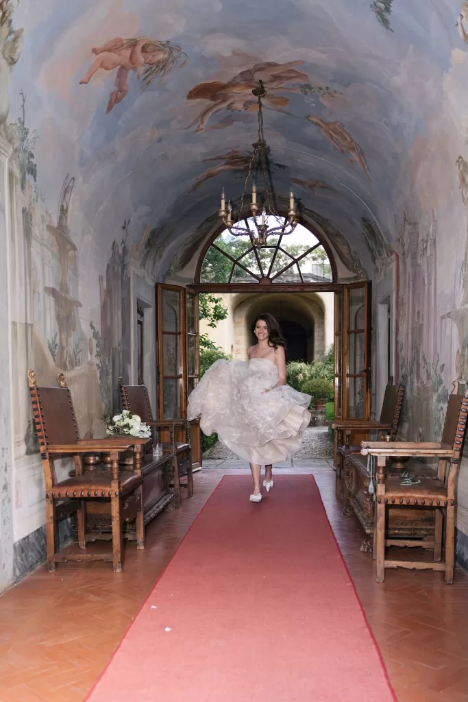 A joyful bride lifts her gown as she walks through a frescoed hallway at Villa Medicea di Lilliano.