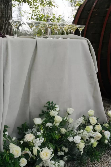 Champagne glasses and white floral arrangements styled on a linen-covered drinks table under the trees at Villa Medicea di Lilliano.
