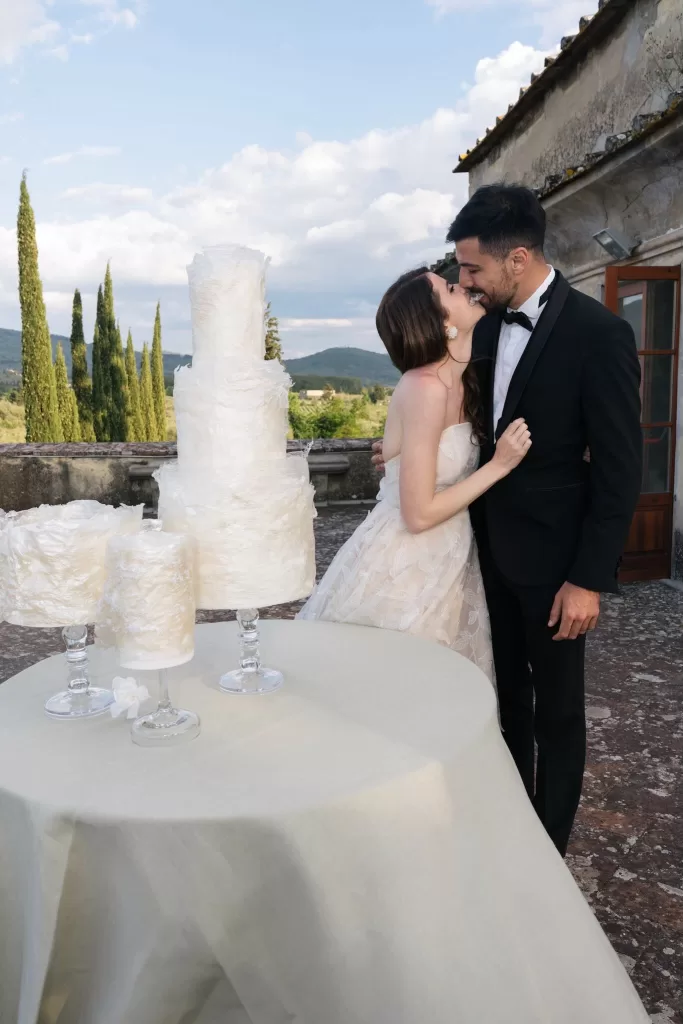 Bride and groom sharing a kiss beside their textured white wedding cakes on the terrace at Villa Medicea di Lilliano.