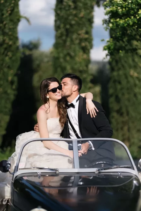 Just married couple in a vintage convertible, sharing a kiss surrounded by Tuscan cypress trees at Villa Medicea di Lilliano.
