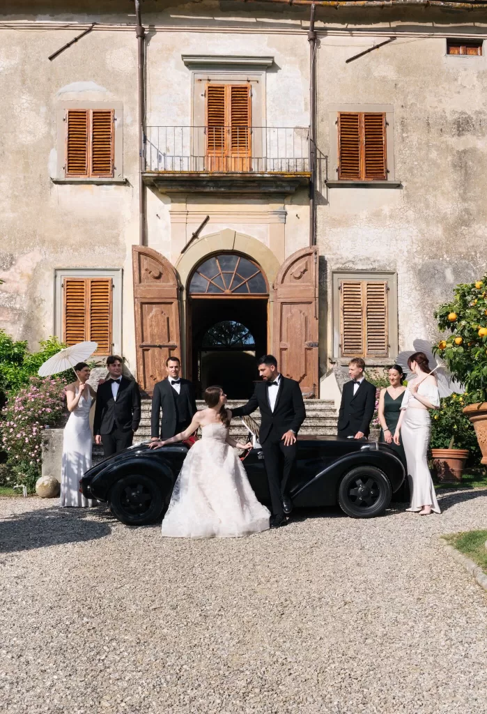 The wedding party poses beside a vintage black car in front of Villa Medicea di Lilliano on a sunlit celebration day.
