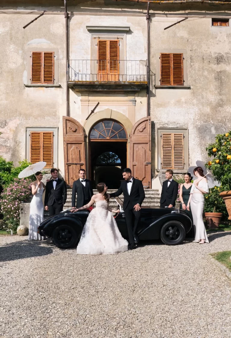 The wedding party poses beside a vintage black car in front of Villa Medicea di Lilliano on a sunlit celebration day.