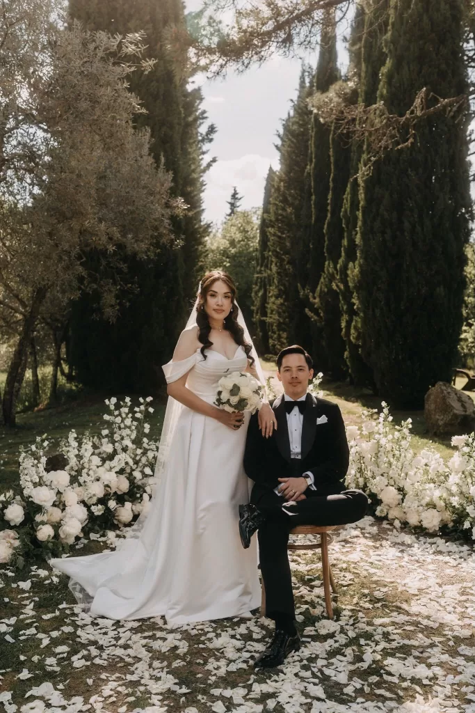 Bride and groom pose in a sunlit floral aisle surrounded by cypress trees at Villa Olimpia