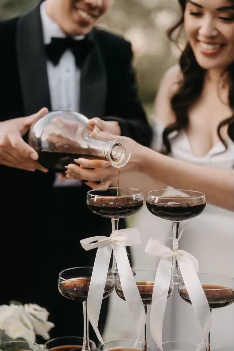 Bride and groom pouring dark cocktails into coupe glasses tied with white ribbons at Villa Olimpia.
