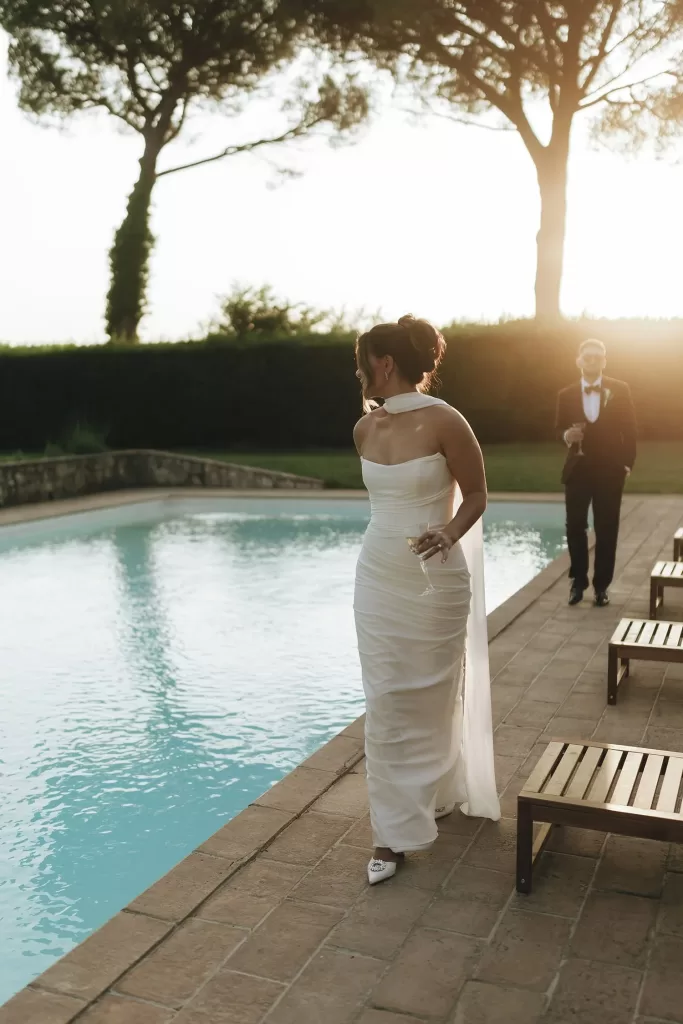 Bride walks along the poolside with a glass of champagne, the groom following behind at Villa Ravano.