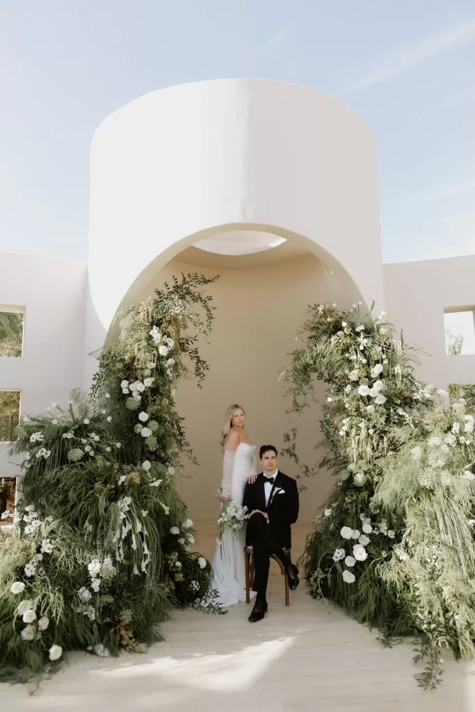 Bride and groom posing under the Baja Luna ceremony arch adorned with greenery and white flowers.