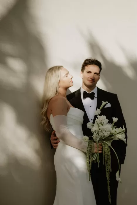 Bride and groom standing against a sunlit wall at Baja Luna, bride holding white bouquet.