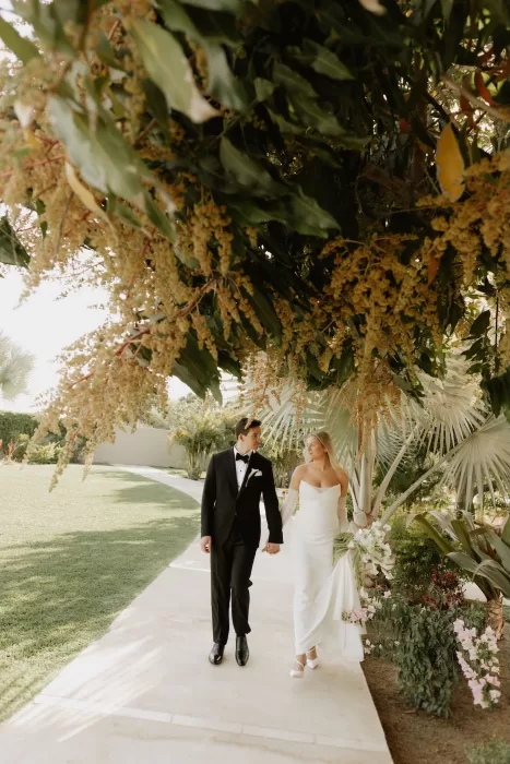 Bride and groom walking hand in hand beneath flowering trees in the gardens of Baja Luna.