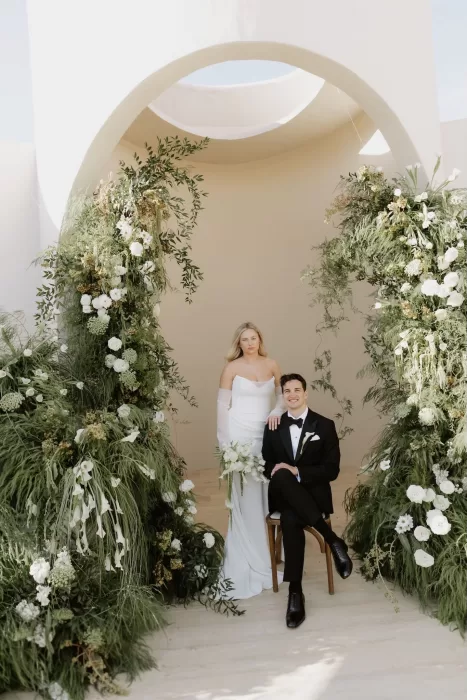 Bride and groom posing beneath a lush green and white floral arch at Baja Luna.