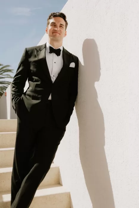 Groom in a classic black tuxedo leaning casually against a white wall at Baja Luna under the bright sun.
