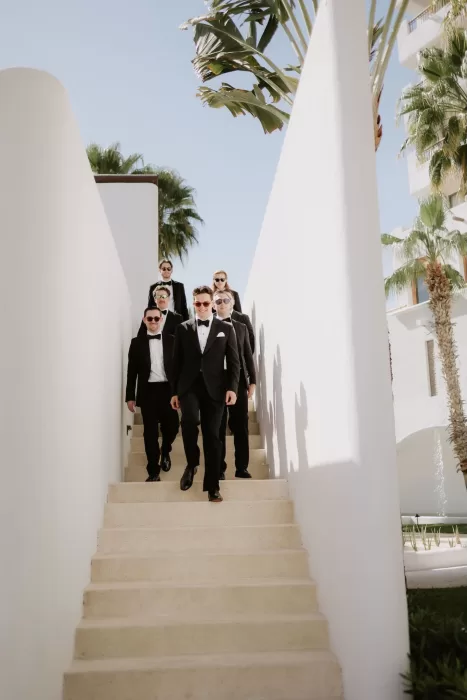 Groomsmen in black tuxedos walking down whitewashed stairs at Baja Luna under palm trees.
