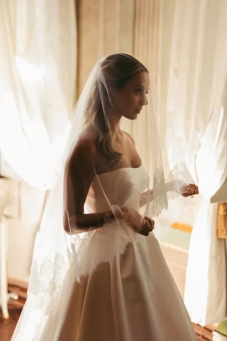 The bride stands in soft window light at Borgo Stomenanno, holding her lace-edged veil.