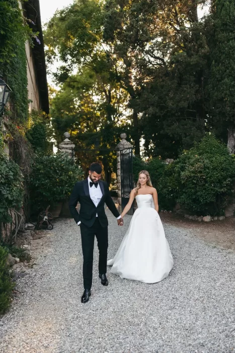 Bride and groom walking hand in hand down a gravel path at Borgo Stomenanno, surrounded by greenery.