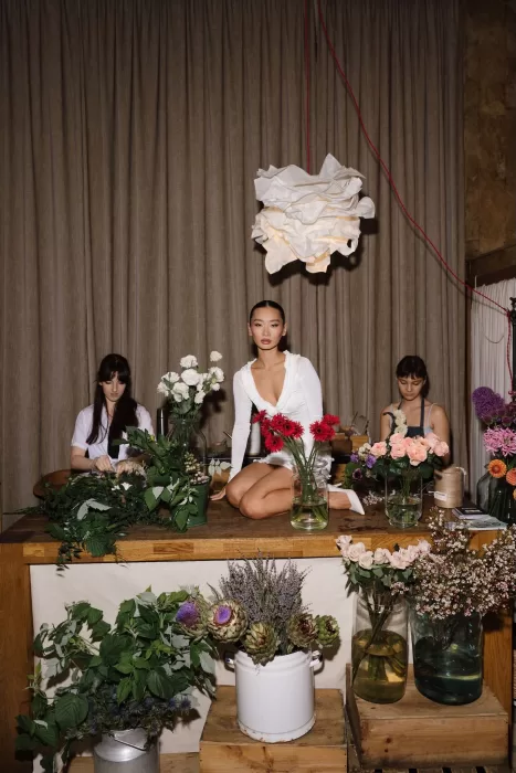 Bride sitting among vibrant flowers on a wooden florist table during wedding evening preparations.
