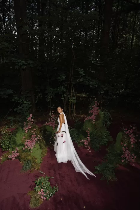 Bride in flowing white gown walking through a woodland aisle lined with moss and pink flowers.
