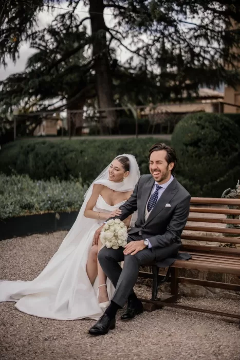 The bride and groom share a joyful moment on a bench in the gardens of Cigarral de la Mercedes, surrounded by lush greenery.