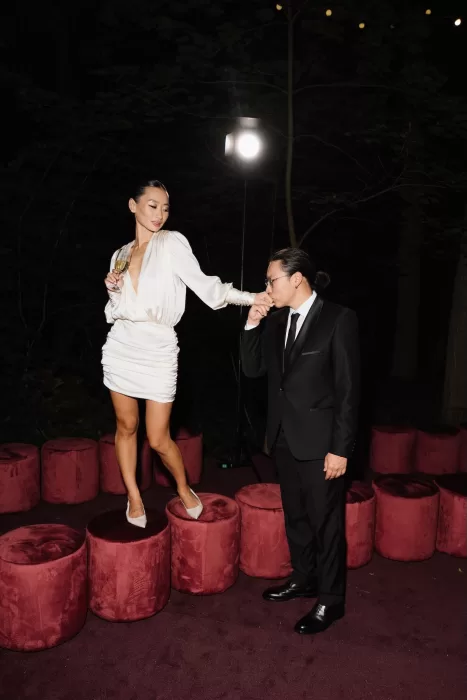 Groom kissing bride’s hand during evening celebration, with bride in short satin dress holding champagne.