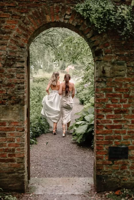Bride walking with a bridesmaid through a rustic brick archway at Middleton Lodge.